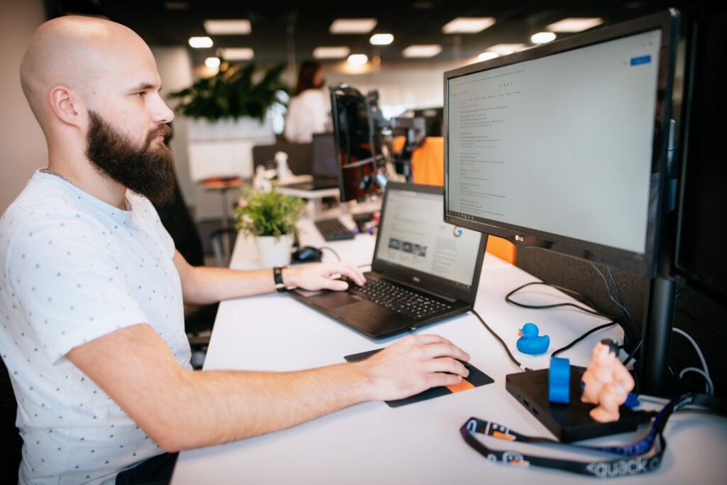 A bearded man sits at a desk in an office, working on a computer with two monitors. A laptop, plant, rubber duck, and office supplies are on the desk. The background shows a modern open workspace.