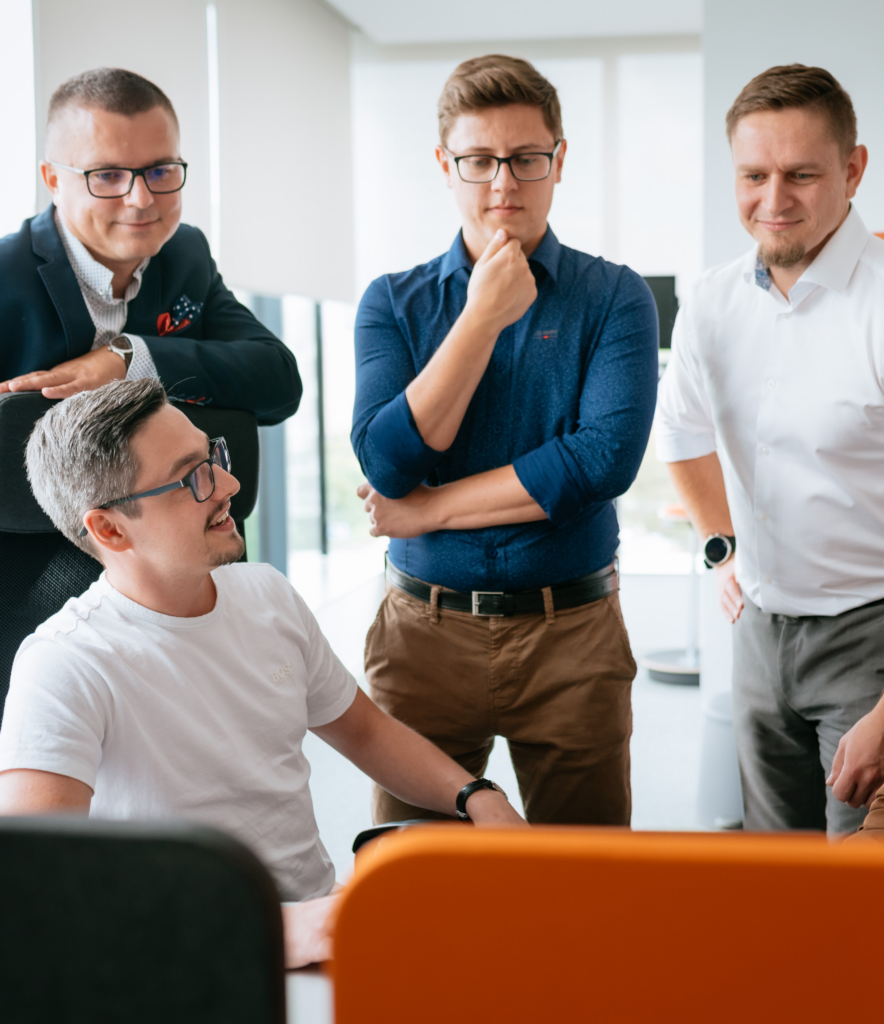 Four men in an office gather around a desk, focused on a computer screen. One man sits and smiles while the other three stand, appearing engaged and thoughtful. Bright windows provide natural light.