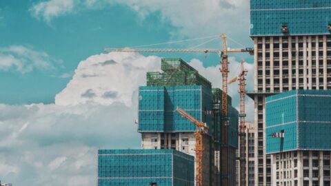Several high-rise buildings under construction with yellow cranes and blue safety netting, set against a bright sky with clouds, highlight the dynamic B2B landscape where companies like Wego Systembaustoffe GmbH are transforming construction material supply through innovative e-commerce solutions.