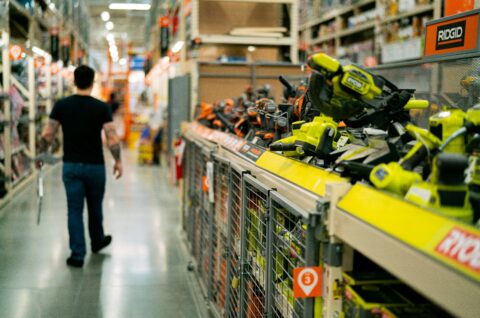 A person walks down a store aisle lined with power tools, including brands like Ryobi and Ridgid, with shelves stacked high and products organized neatly—a scene reflecting the E-commerce Transformation that companies like Wego Systembaustoffe GmbH are bringing to the B2B marketplace.